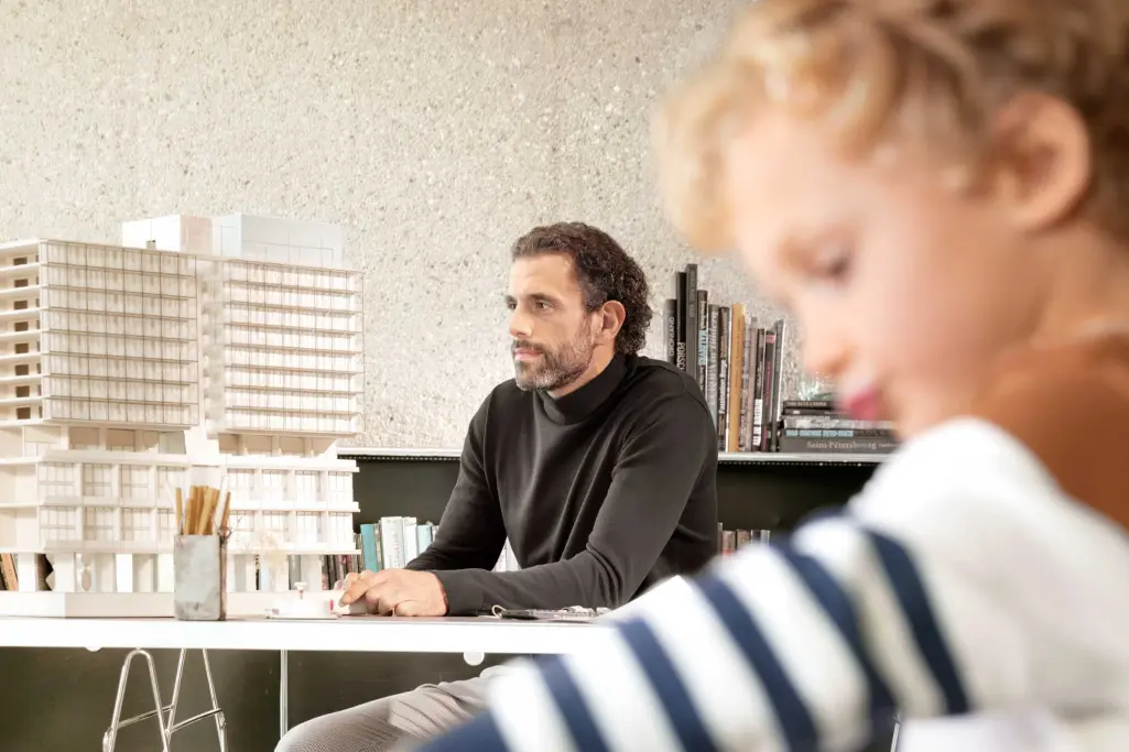 Foto: vorne spielendes Kind in Unschärfe, im Hintergrund Vater, Architekt sitzend am Schreibtisch.