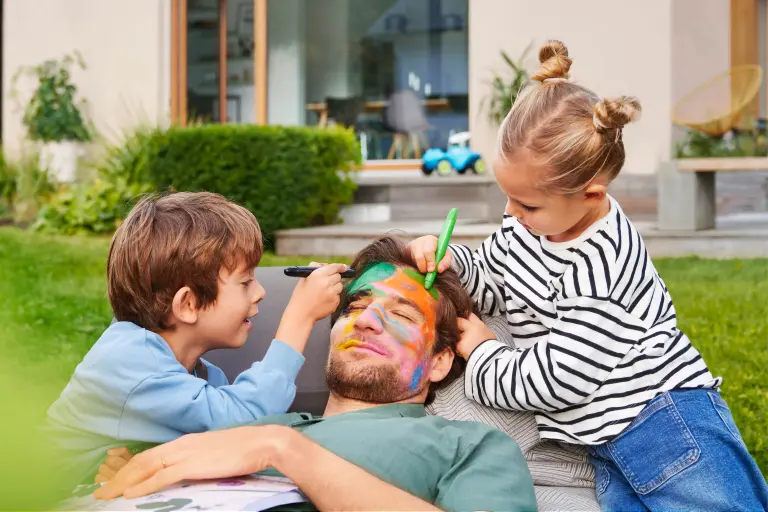 Kinder bemalen vergnügt das Gesicht ihres Vaters, im Hintergrund das frisch sanierte Einfamilienhaus