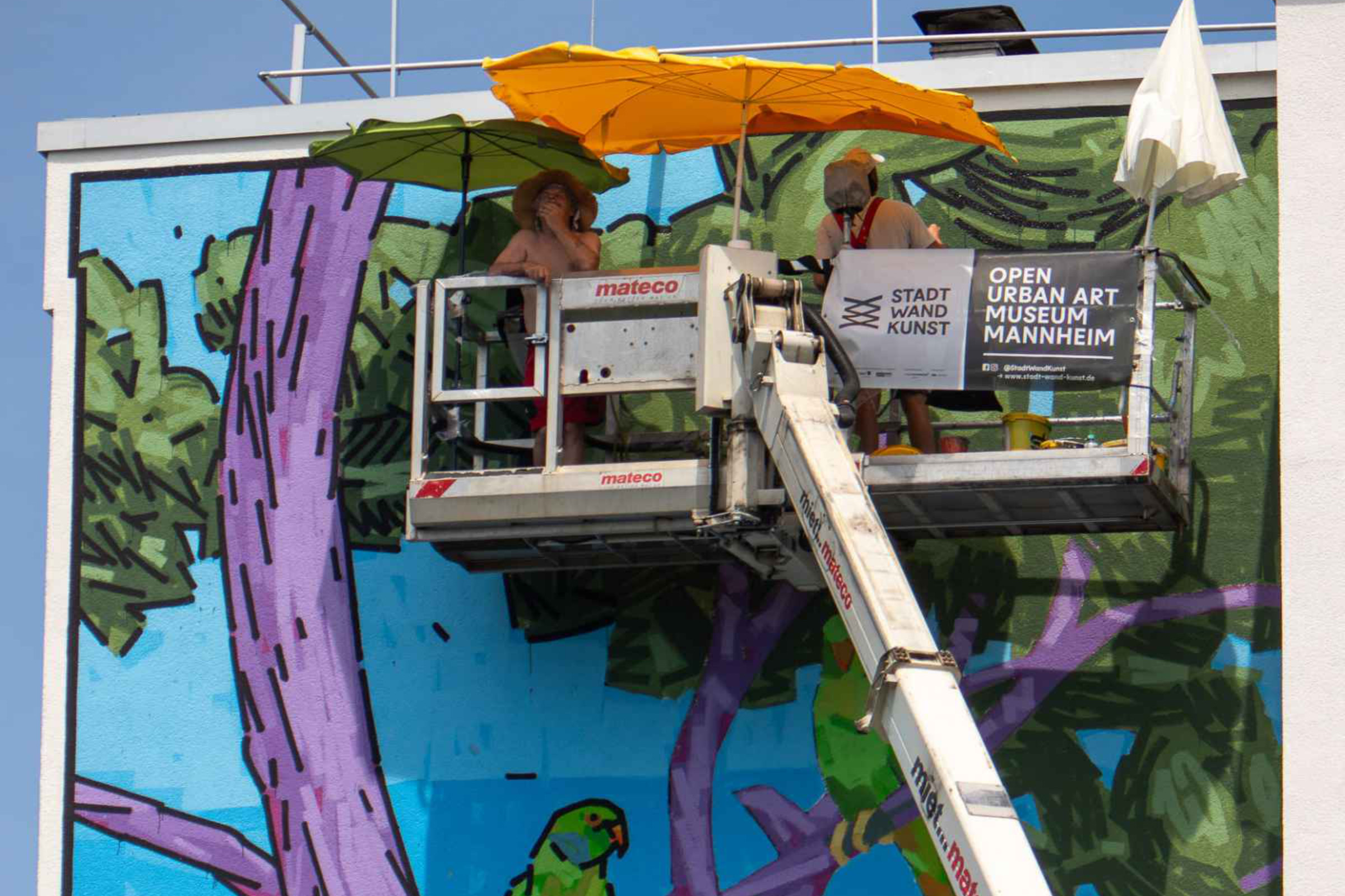 Auf Hebebühne arbeiten Künstler unter Sonnenschutz an dem Kunstwerk an einer Hochhaus-Fassade. – Das Mural „In Contemplation“ von vom Künstler TAXIS in seiner Entstehung an der Fassade eines Wohn-Hochhauses im Mannheimer Stadtteil Lindenhof. 