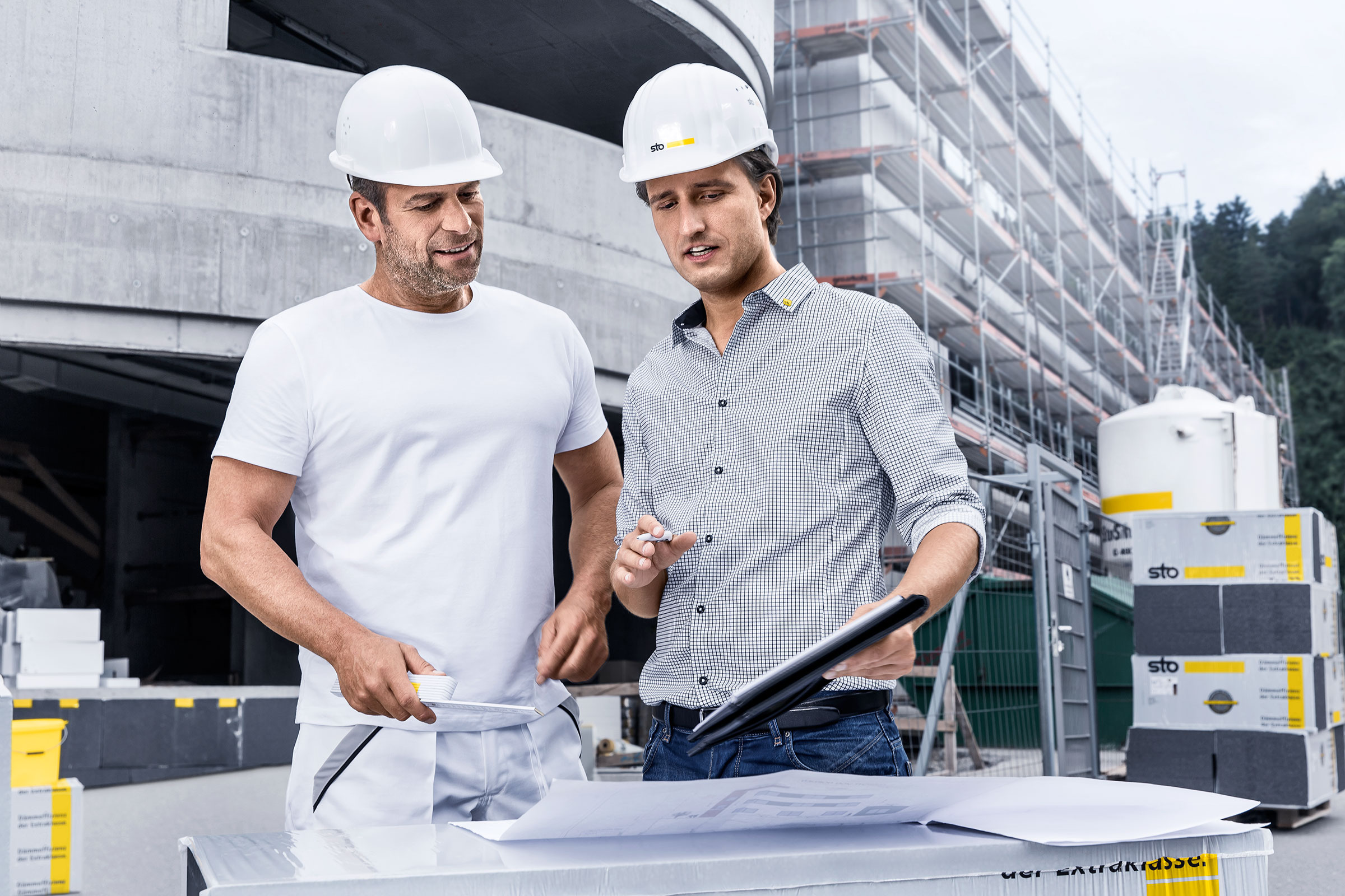 Zwei Bauprofis mit Schutzhelmen besprechen Baupläne auf Baustelle mit Gerüst und Baustoffen