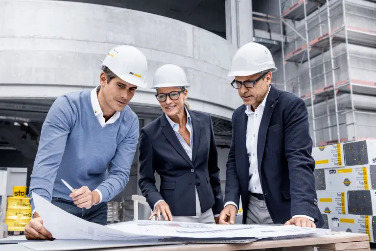 Beratungsfoto auf der Sto-Baustelle: Projektmanager und Architekten, zwei Herren, in der Mitte eine Dame. Alle tragen weiße Helme